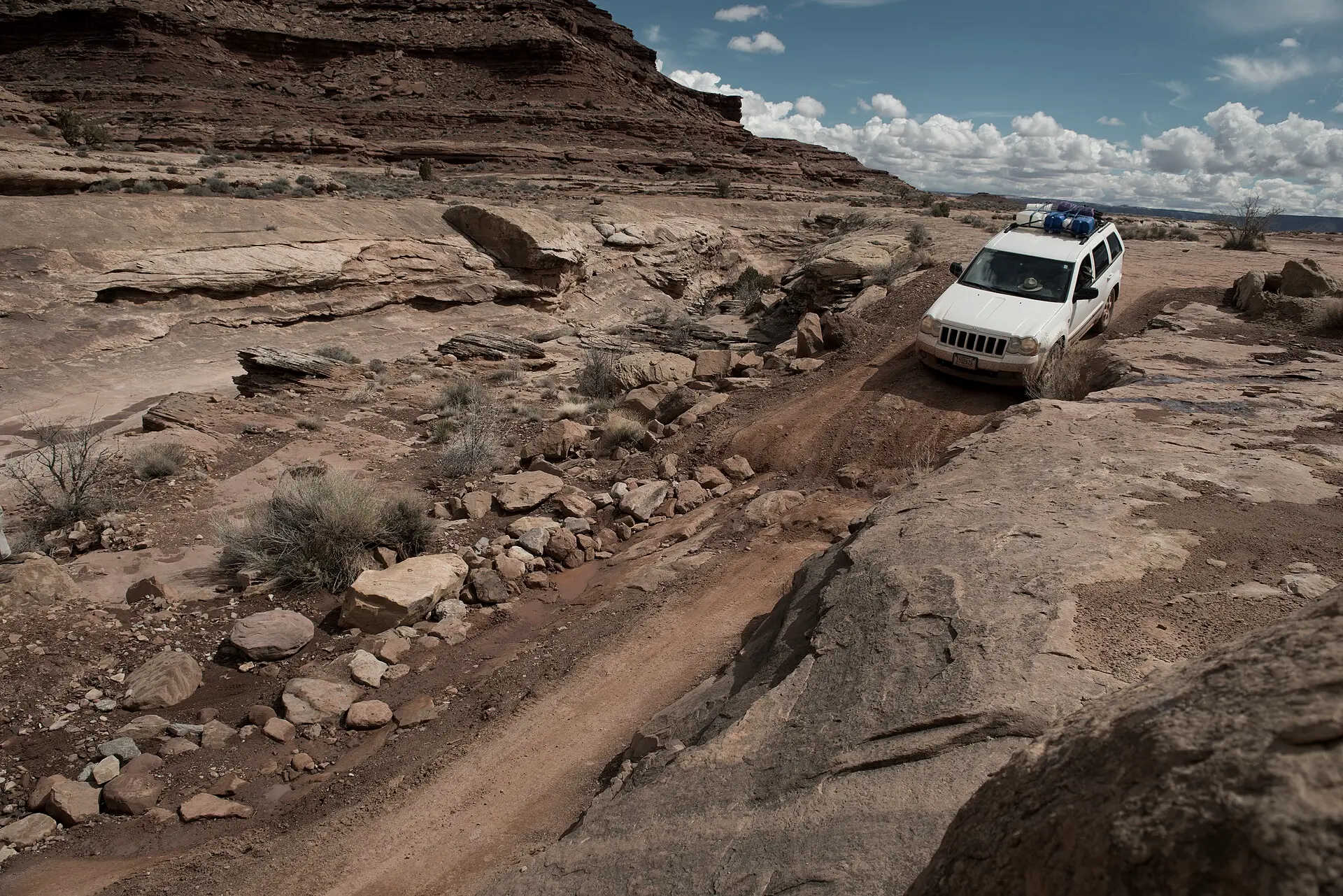 A white 4x4 with a loaded roof rack climbing a rocky red-sand trail beneath a sandstone mesa.